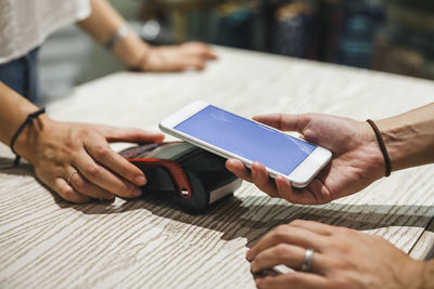 Cropped image of female customer making contactless payment with smart phone to owner in clothing store