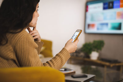 Side view of young woman controlling tv through mobile app at home