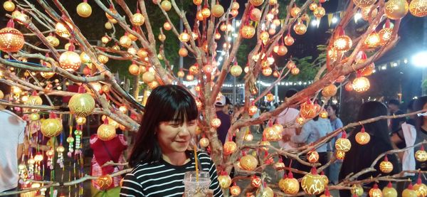 Smiling woman drinking juice while standing against illuminated tree at night