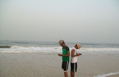 Friends using mobile phone while standing at beach against sky