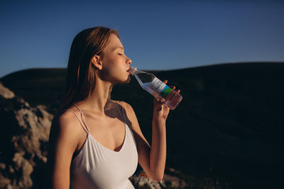 Young woman drinking water from bottle against sky