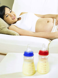 Pregnant woman looking at milk bottles while lying down on sofa at home