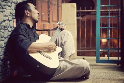 Man playing guitar while sitting by wall