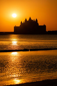 Silhouette buildings in sea against orange sky during sunset