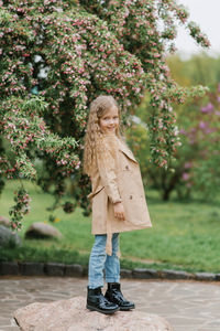 Happy girl child with long wavy brown hair stands on a stone in spring in a blooming park 