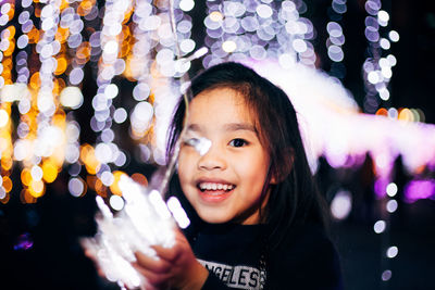Close-up portrait of smiling girl holding illuminated string lights at night
