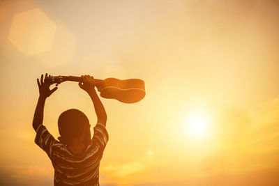 Rear view of boy with guitar standing against sky during sunset