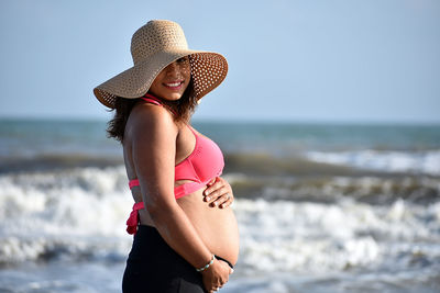 Side view of pregnant young woman in hat standing at beach against sky