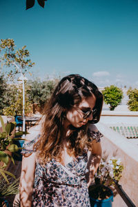 Young woman looking down while standing against plants against clear sky