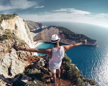 Rear view of man with arms outstretched standing by sea on cliff during sunny day