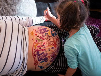 Girl painting pregnant mother belly on sofa at home