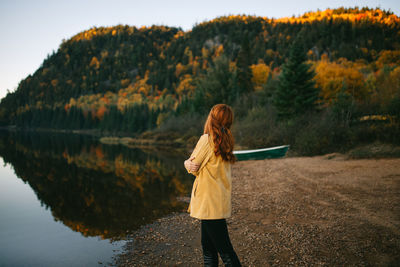 Side view of unrecognizable young female traveler with ginger hair admiring amazing autumn landscape while standing on shore of calm lake surrounded by lush forest during trip in canada