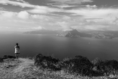 Rear view of woman looking at sea while standing on mountain against cloudy sky