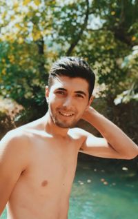 Portrait of shirtless young man smiling against lake in forest