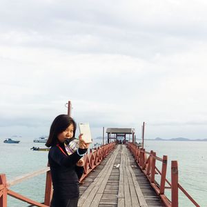 Young woman taking selfie while standing on pier against cloudy sky