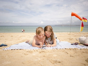 Siblings sharing smart phone while lying on towel at beach against sky