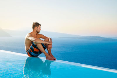 Young man sitting at poolside against sea