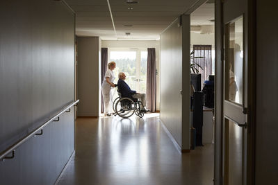 Full length side view of female nurse pushing senior man on wheelchair at hospital corridor