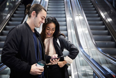 Smiling man showing smart phone to female friend while standing on escalator at underground station