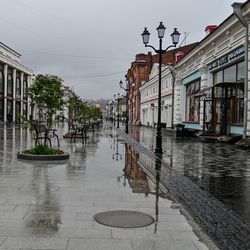 Wet street by buildings against sky during rainy season