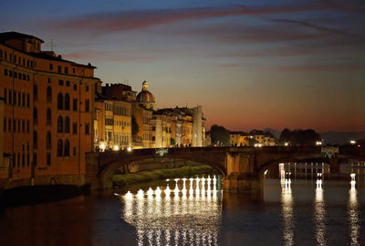 Bridge over river by illuminated buildings against sky during sunset