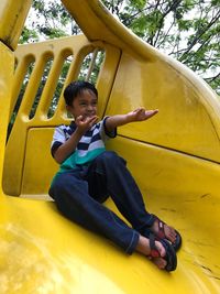 Boy gesturing horn sign while sitting on yellow slide at playground