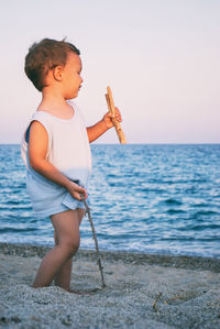 Boy holding sticks standing at beach against clear sky