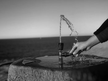 Cropped hand at drinking fountain by sea against clear sky