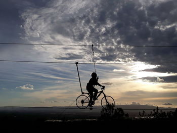 Silhouette man riding bicycle on cable against cloudy sky during sunset