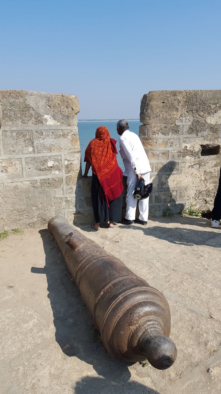 built structure, building exterior, architecture, clear sky, sunlight, day, outdoors, full length, beach, blue, house, sand, sky, childhood, copy space, person, stone wall, stone - object, rear view