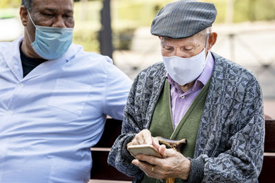 Senior man using smart phone wearing protective face mask sitting with mature male on bench during covid-19