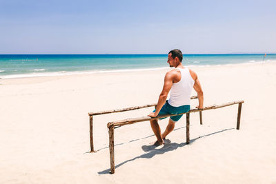 Rear view of man sitting on parallel bars at beach during sunny day