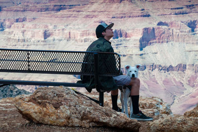 Side view of teenage boy sitting on bench with dog against rock formations