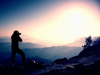 Amateur photographer takes photos with camera on peak of rock. dreamy landscape, pink misty sunrise