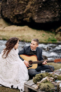 Man playing guitar while sitting with woman near river