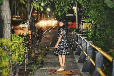 Full length portrait of young woman standing on sidewalk in city at night