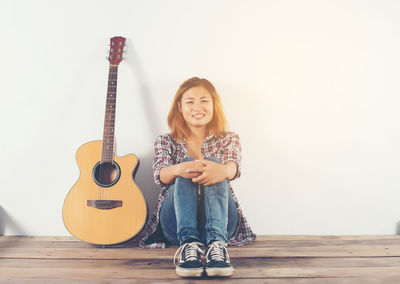 Portrait of smiling young woman sitting by guitar against white wall