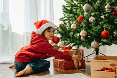 Charming boy in a santa hat takes out christmas gifts from under the new year tree and laughs