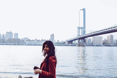Portrait of young woman standing on bridge over river against clear sky