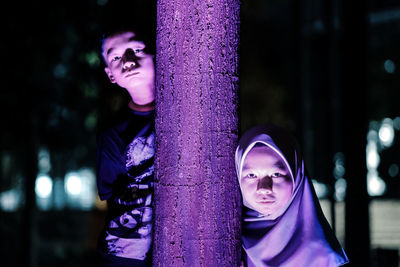 Portrait of siblings standing by tree trunk at night