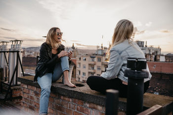 Cheerful women talking while sitting on building terrace against sky