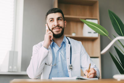 A young doctor of oriental appearance is talking on the phone, sitting at the table 