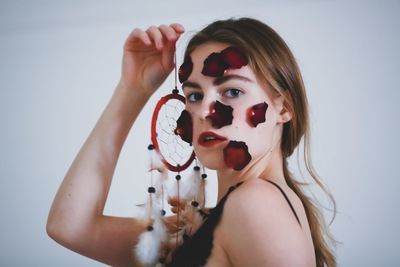 Portrait of woman with rose petals on face holding dreamcatcher against white background