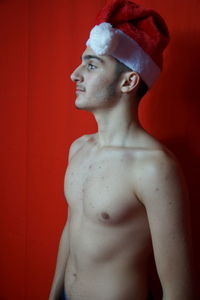 Shirtless young man wearing santa hat standing against red wall