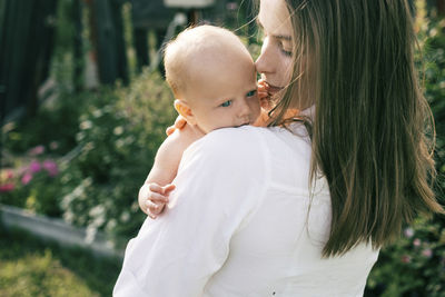 Rear view of mother carrying cute daughter while standing against plants in park