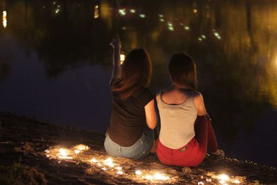 Rear view of female friends sitting by illuminated tea light candles at lakeshore during night
