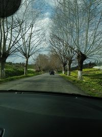 Road by bare trees against sky seen through car windshield