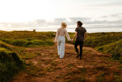 Back view of unrecognizable man and woman in casual clothes holding hands and walking against cloudy sky in evening in countryside of aviles, spain