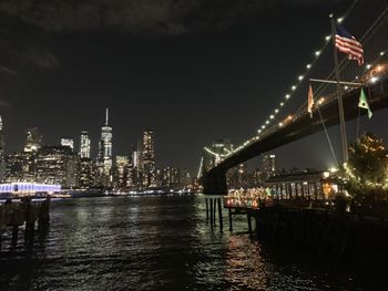 Illuminated bridge over river by buildings against sky at night