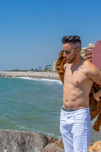 Young man wearing sunglasses standing on beach against clear sky